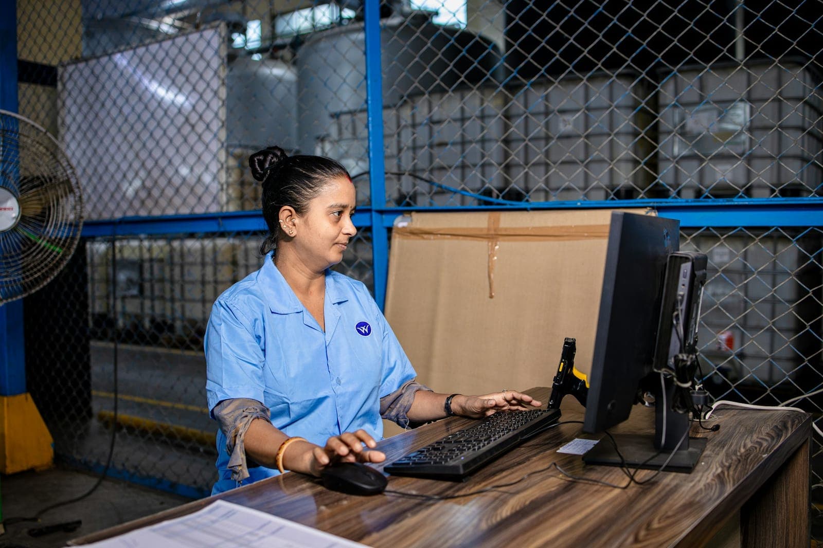 Funcionária de uniforme azul digita em um computador numa mesa dentro de um depósito