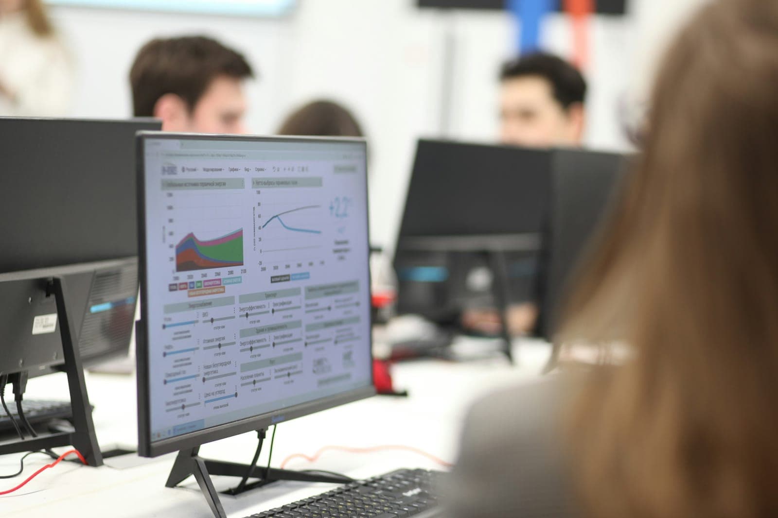 Monitor com gráficos em primeiro plano numa sala onde várias pessoas trabalham em computadores