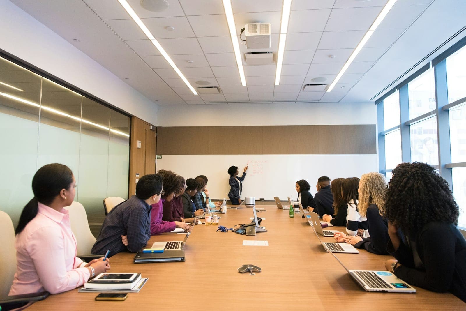 Mulher de pé escreve num quadro branco diante de uma equipe sentada em volta de uma mesa longa de reunião com notebooks