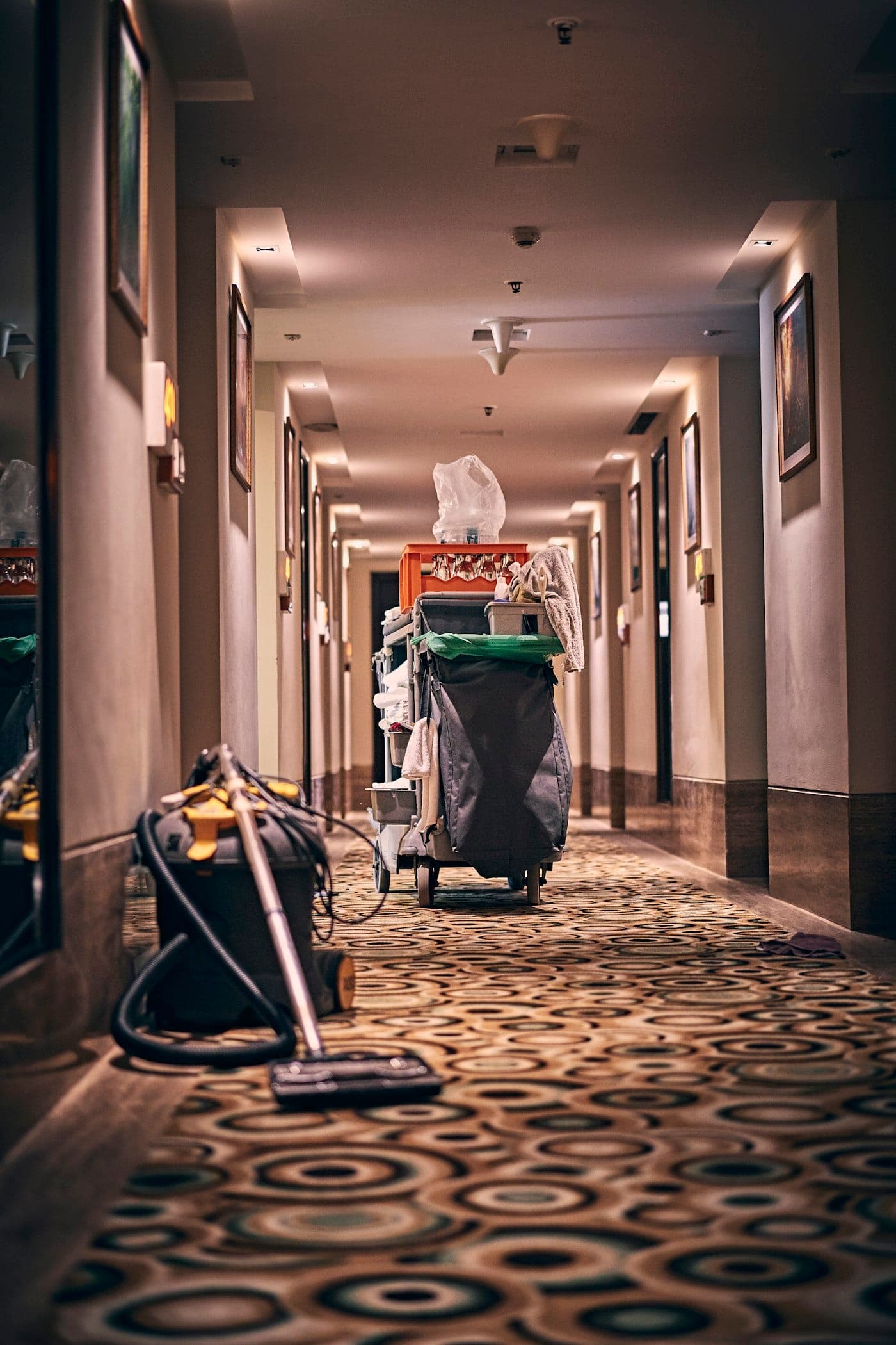 Corredor de hotel com carpete estampado, um carrinho de governança carregado de enxoval ao fundo e um aspirador de pó em primeiro plano